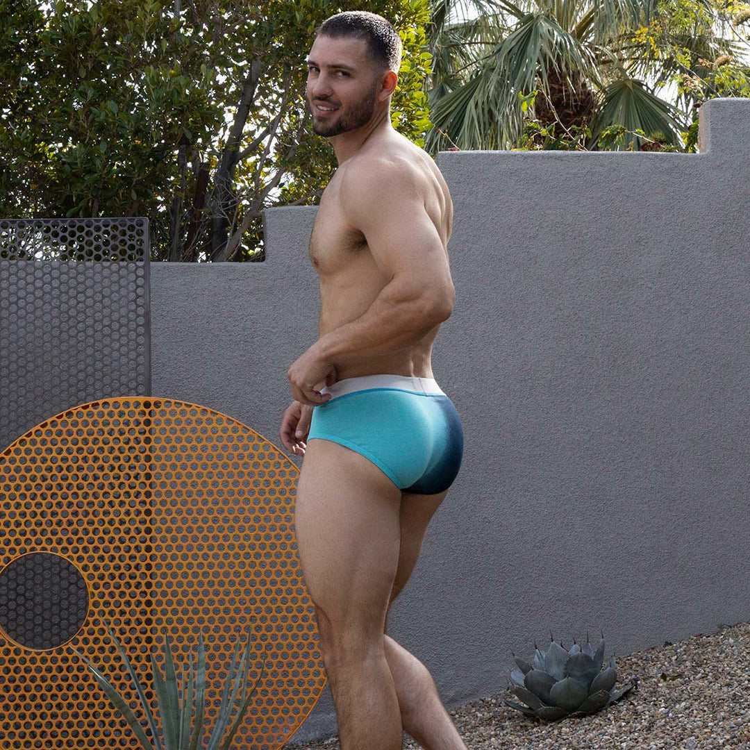 Man wearing a blue swimsuit standing outdoors with a gray wall and plants in the background