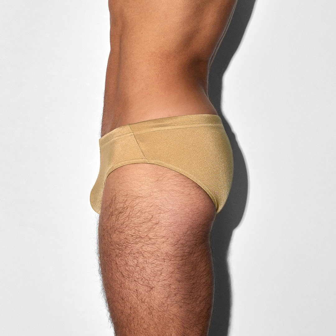 A side profile view of a man's lower torso and upper leg wearing a pair of low-rise, light tan/beige swim briefs against a light gray background.
