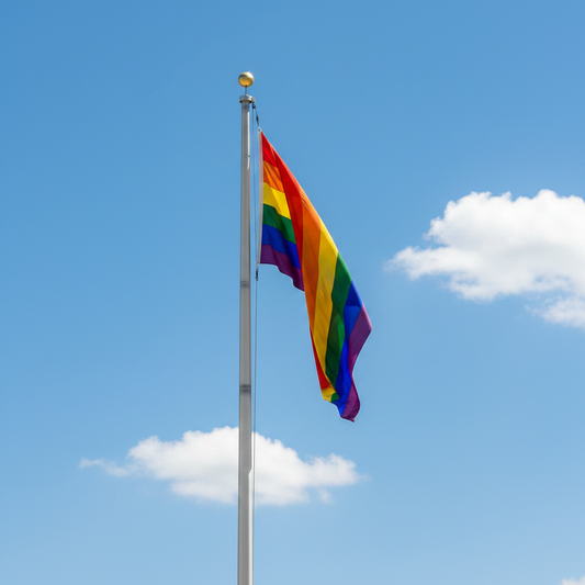 A rainbow pride flag waving on a silver flagpole against a bright blue sky with white clouds.