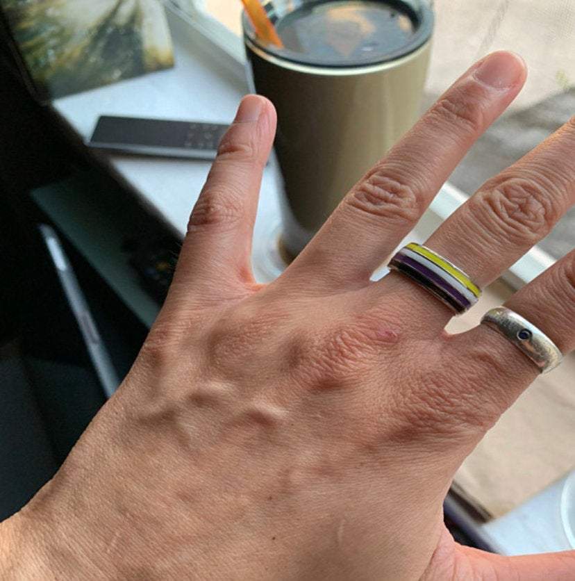 Hand wearing a nonbinary striped ring on a blurred background of a table with a cup and phone.