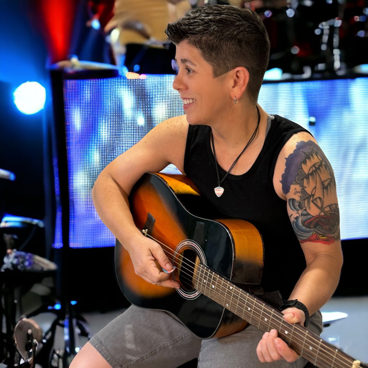 Person playing an acoustic guitar on a stage wearing a trans pride guitar pick necklace with lights and equipment in the background