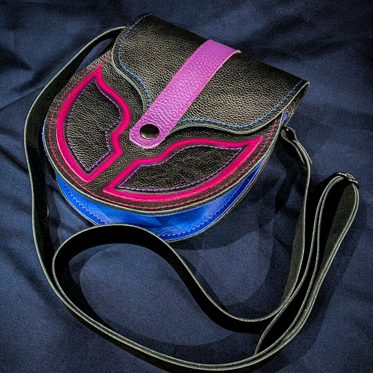 A black textured leather sporran with a bright pink, purple and blue bi pride design on the front flap, resting on a dark navy blue fabric background
