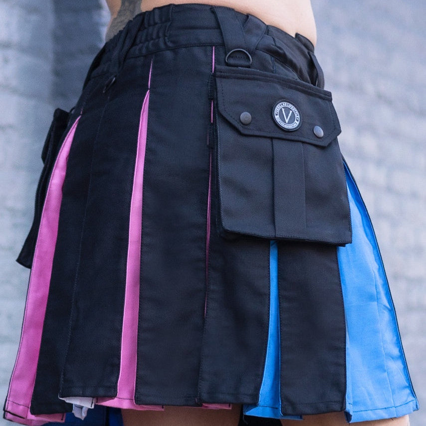 A close-up of a person wearing a black pleated kilt with vertical panels in bright pink, blue, and white to represent the transgender pride flag, plus a flap pocket with a circular 'V' logo. The photo is cropped from the waist to the boots against a gray wall.