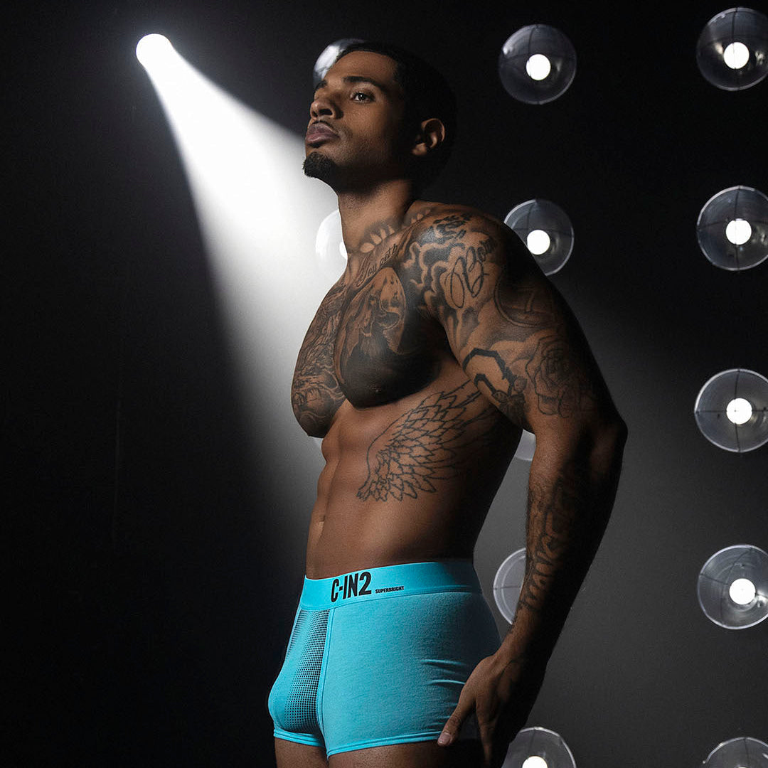 A tattooed male model wearing bright turquoise blue boxer briefs with a black waistband logo, posed against a dark studio background with dramatic spotlights and circular light fixtures.