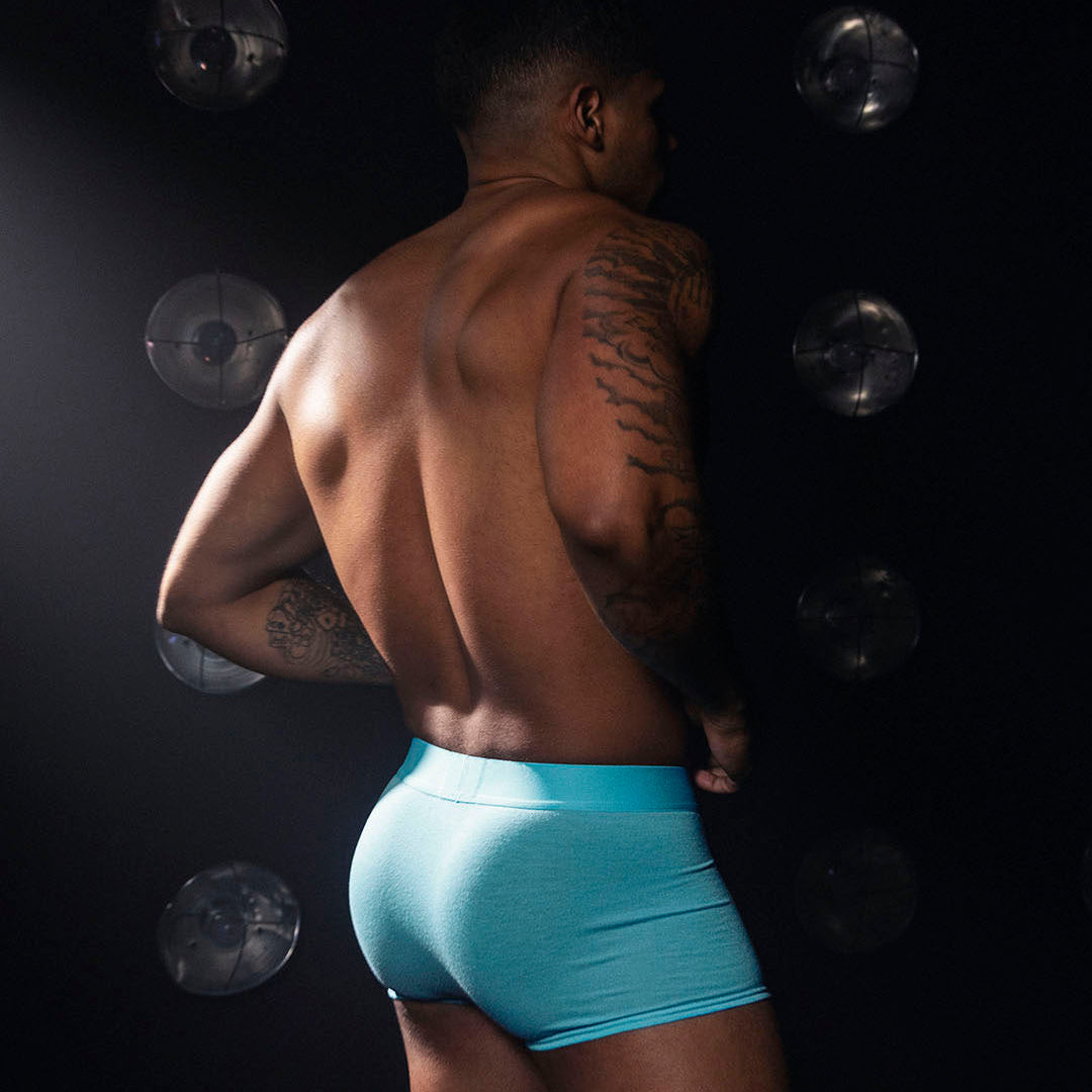 A man shown from the back wearing bright aqua blue boxer briefs against a dark studio background with circular light effects.