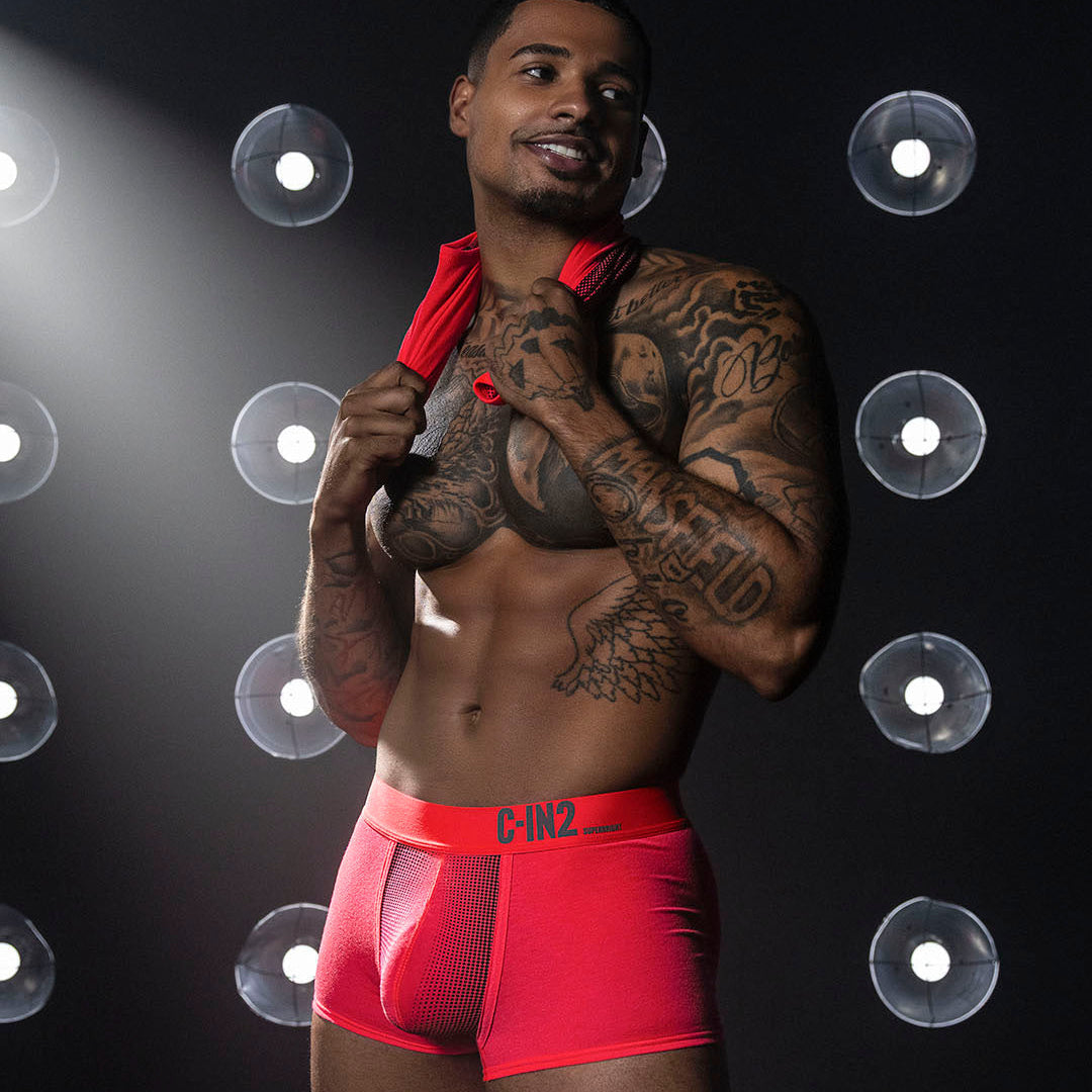 A tattooed male model wearing bright red boxer briefs with a red towel over his shoulders, posed against a dark studio background with circular lights.