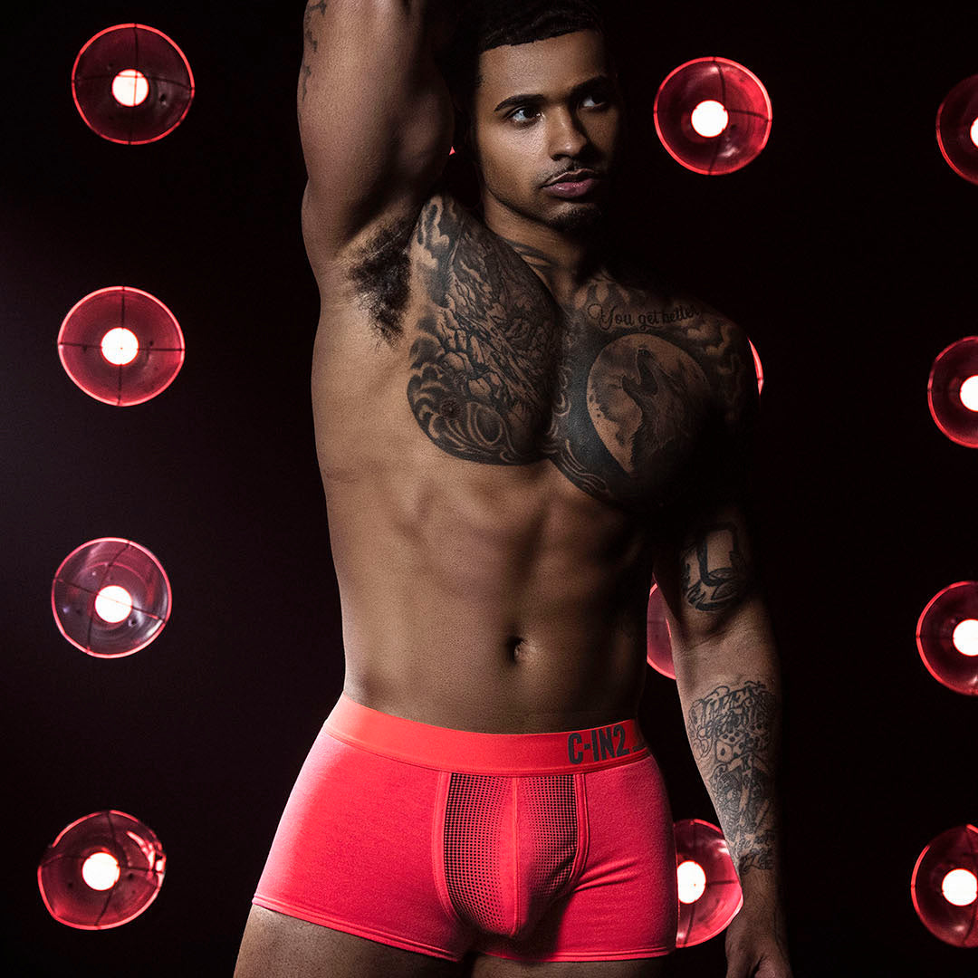 A shirtless male model with tattoos wearing bright red boxer briefs, posed against a dark background with circular red lights.