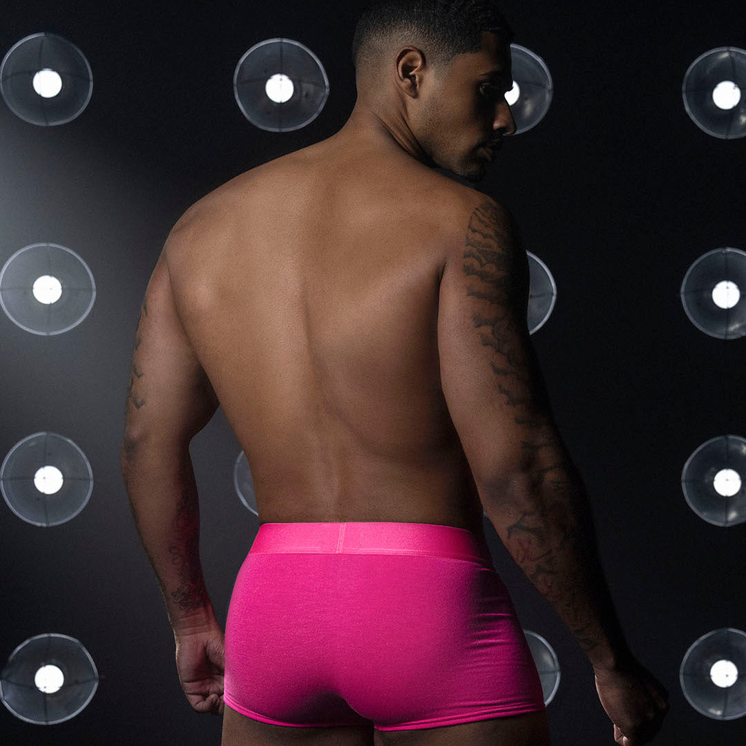 A man shown from the back wearing bright hot pink boxer briefs, standing against a dark background with circular studio lights.