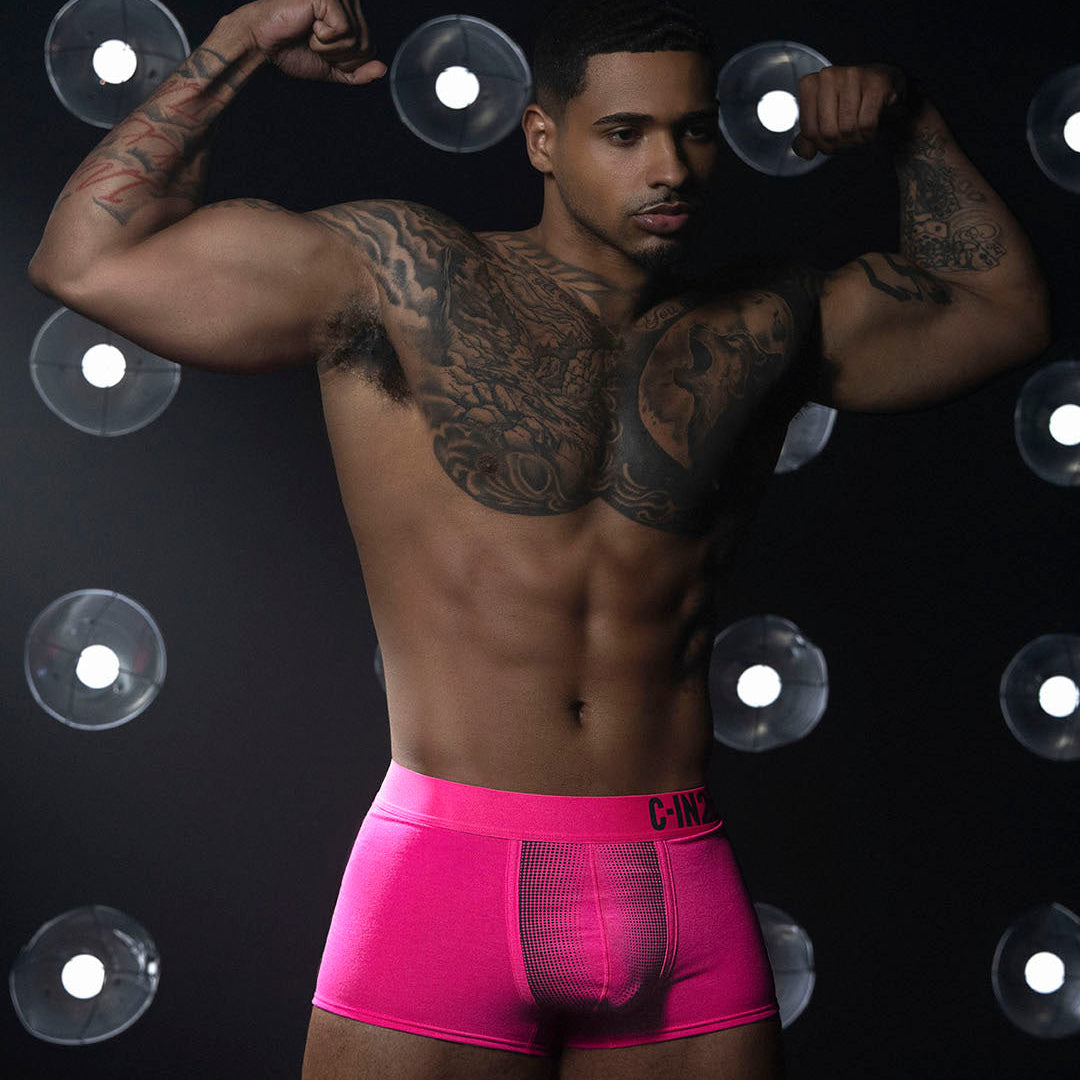A muscular man with tattoos poses in bright pink boxer briefs against a black background with circular light fixtures.