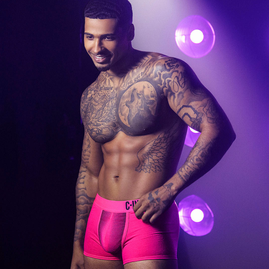 A tattooed male model wearing bright hot pink boxer briefs with a black waistband logo, posed against a dark background with purple stage lighting.