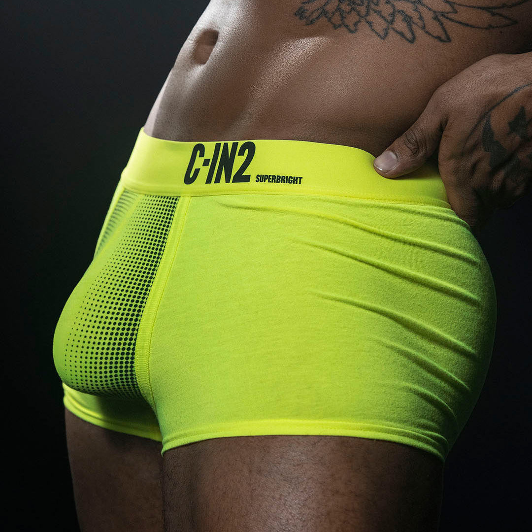 A close-up of a model wearing bright neon yellow-green boxer briefs with a black C-IN2 SUPERBRIGHT logo on the waistband, set against a dark background.