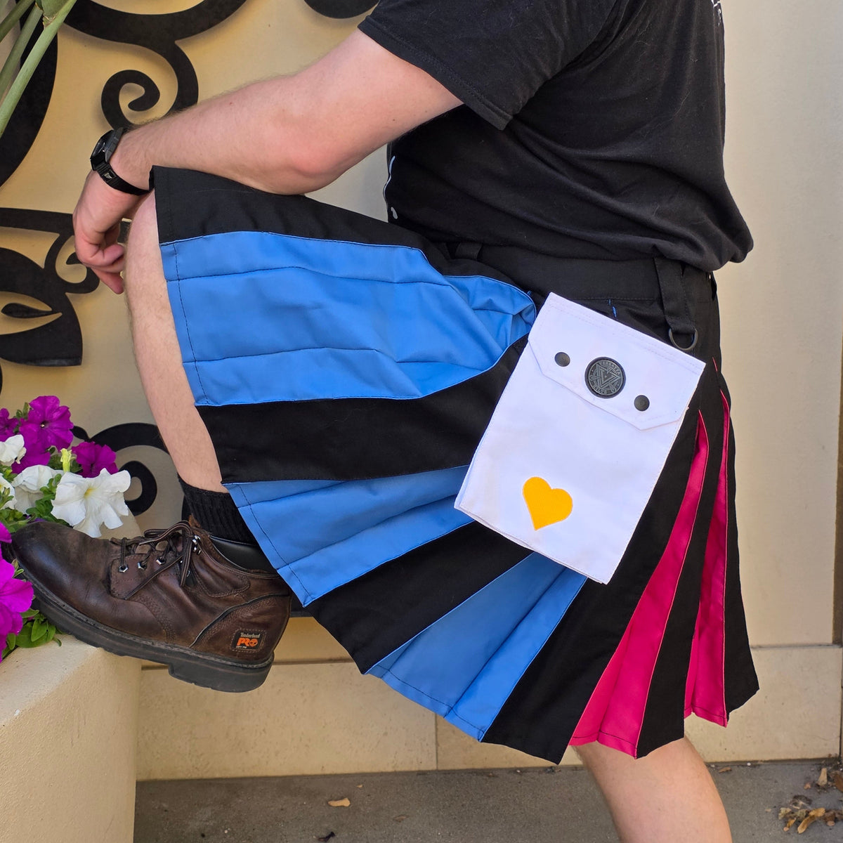 Person wearing a colorful kilt with a white cargo pocket featuring a black circle, gray square, and yellow heart on a patterned floor.