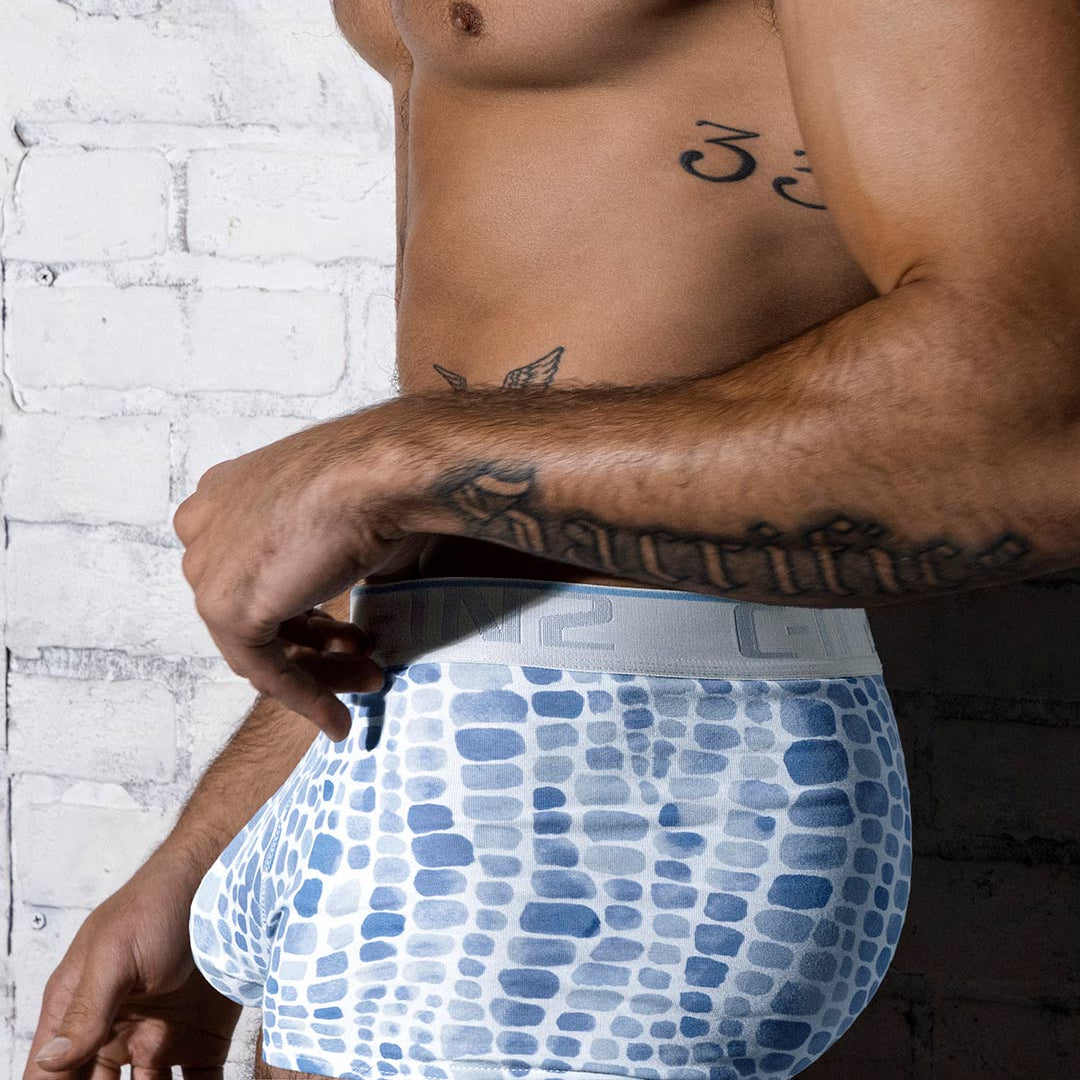 A close-up of a man's torso wearing blue and white patterned boxer briefs against a white brick wall background.