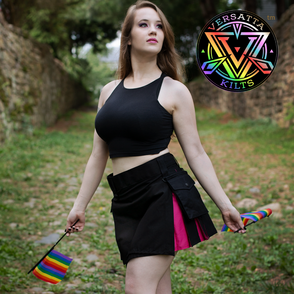 A woman stands outdoors on a grassy path wearing a black crop top and a black kilt with bi pride colored layered flaps. She holds small rainbow pride flags, and a circular Versatta Kilts logo appears in the top right corner.