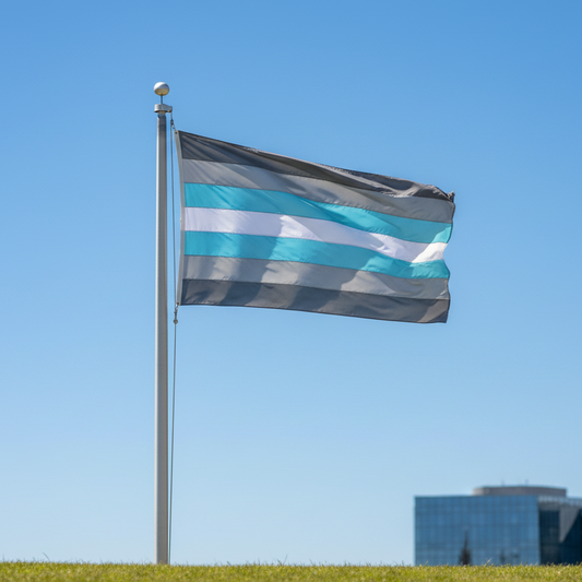A demiboy pride flag with horizontal stripes of dark gray, light gray, white, and light blue waving on a silver flagpole outdoors against a clear blue sky, with a grassy field in the foreground and modern glass buildings in the distance.