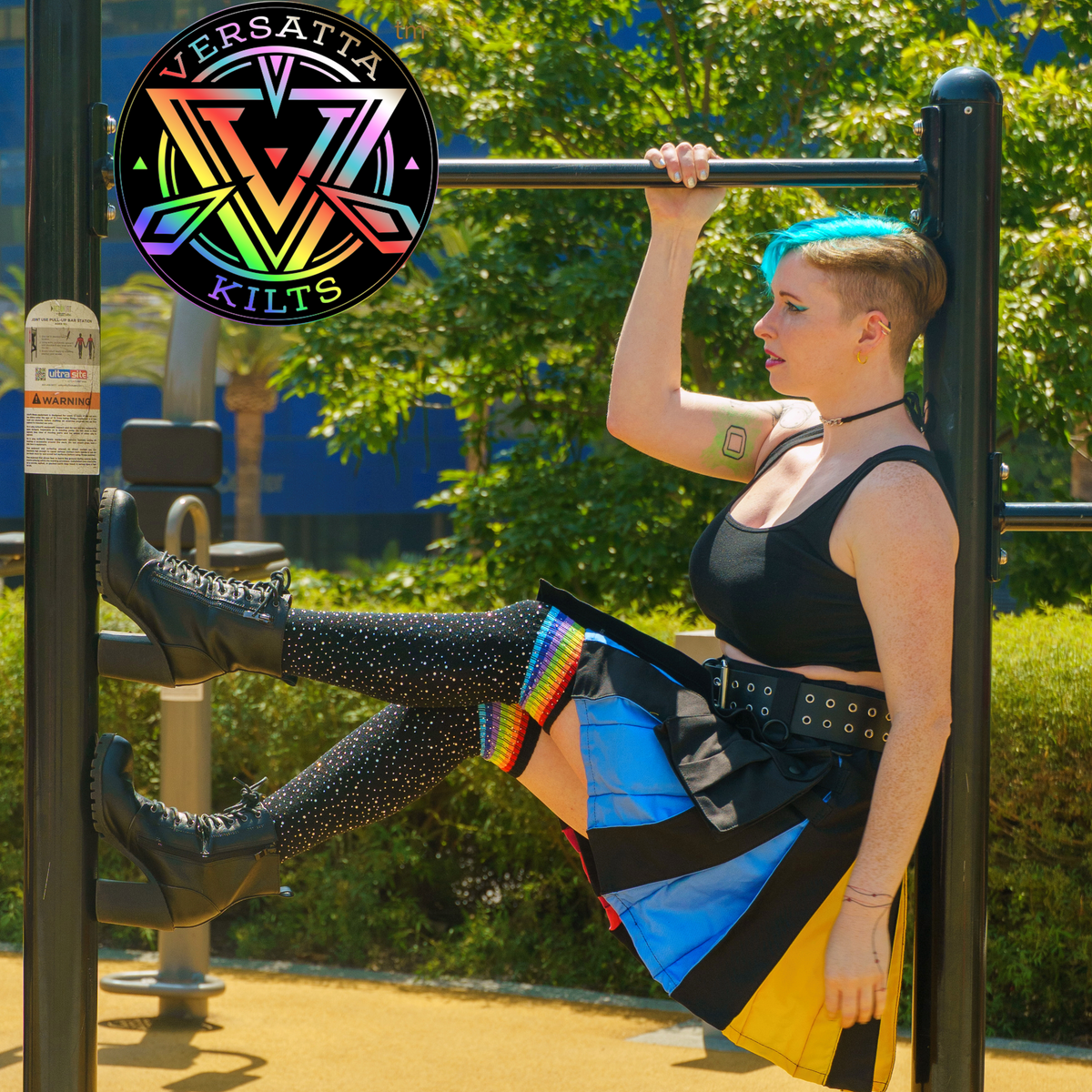 A person with a buzz cut and bright blue hair hangs from a pull-up bar in an outdoor park setting, wearing a black crop top, a colorful kilt with pansexual pride colored trim, black studded tights, and black platform boots. A circular Versatta Kilts logo is overlaid in the top left.