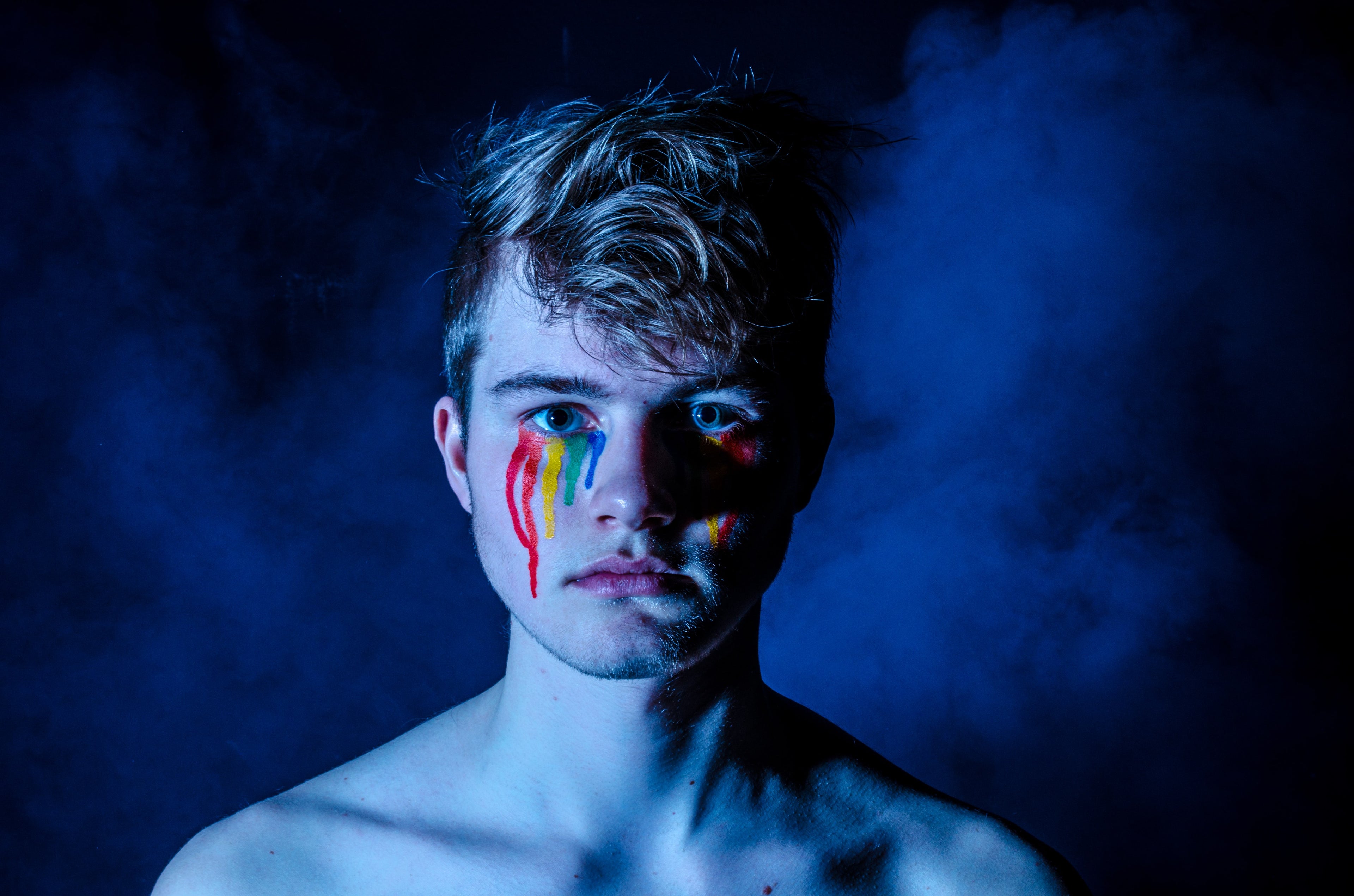 Masculine presenting individual with rainbow colored tears on in a smokey blue background 