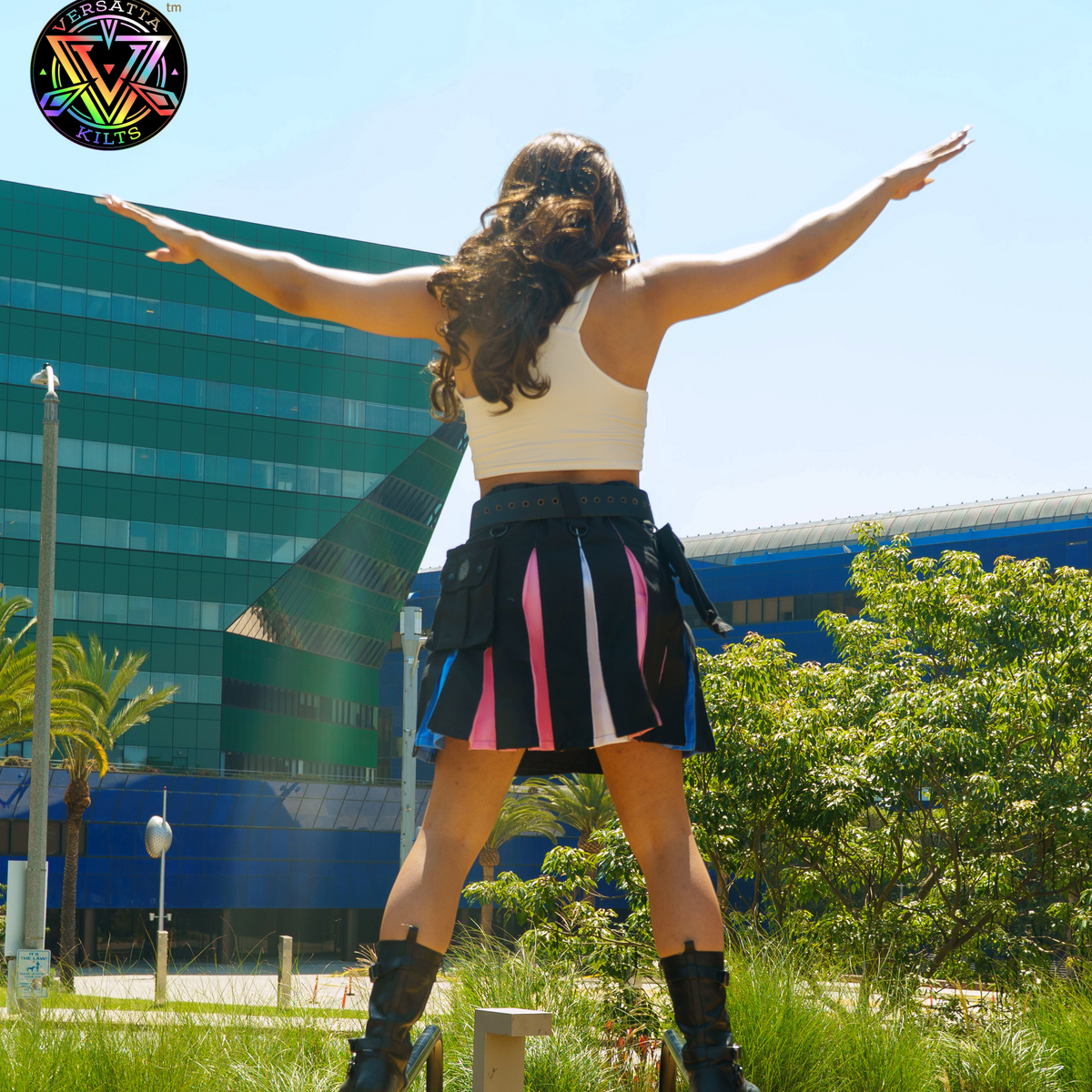 Person photographed from behind showing the kilt they are wearing that is inspired by the trans pride flag colors against traditional black, they have their arms out, set against a modern building and trees