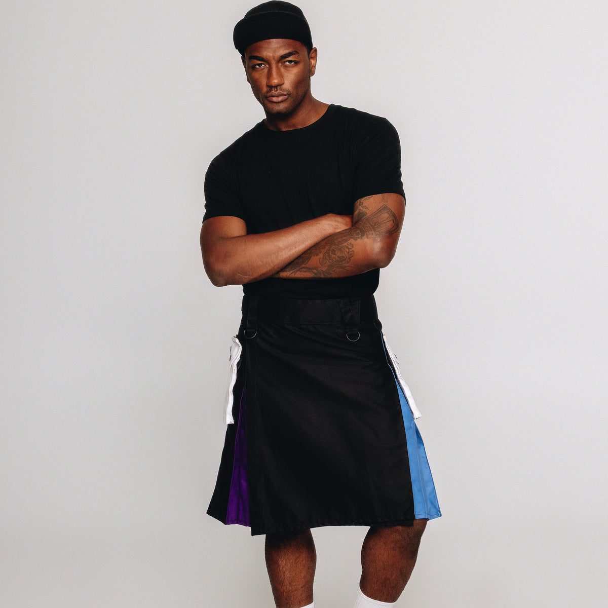 A man modeling a black short-sleeve t-shirt, a black kilt with purple and blue side panels, white socks, and black shoes, standing against a light gray studio background.