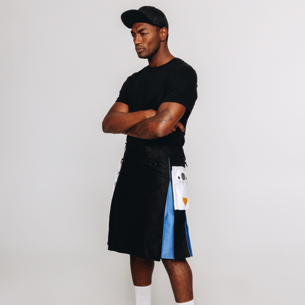 Man wearing a black t-shirt, black and blue kilt, white socks, and black shoes on a white background