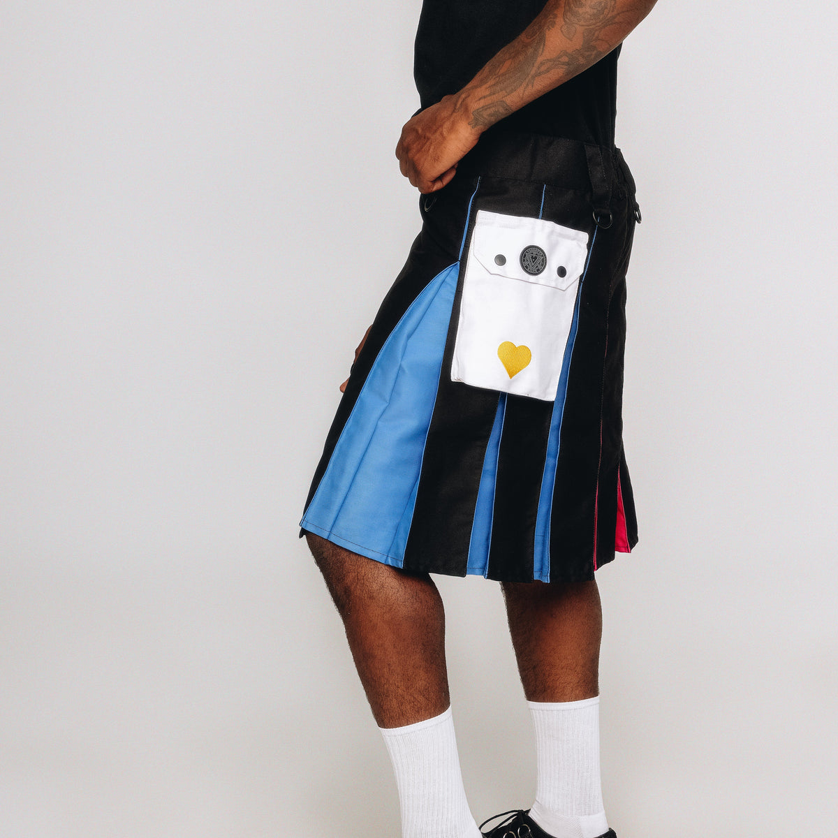 Person wearing a black and blue pleated kilt with a white cargo pocket featuring a yellow heart on a plain background