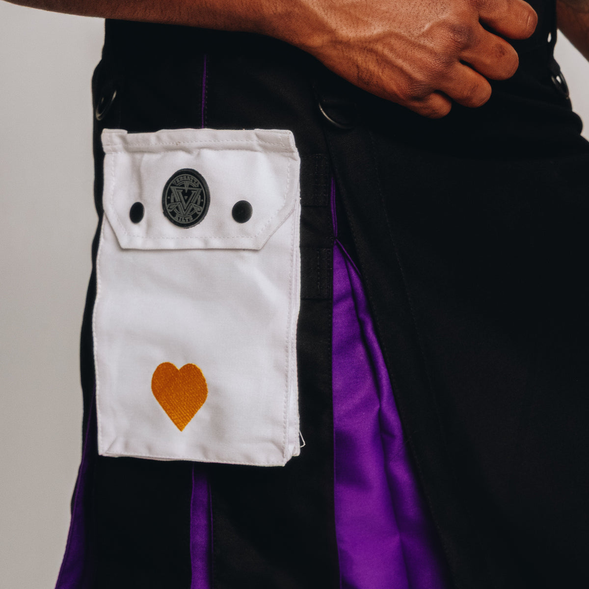 Person wearing a black and purple kilt with a white pocket featuring a heart design on a plain background