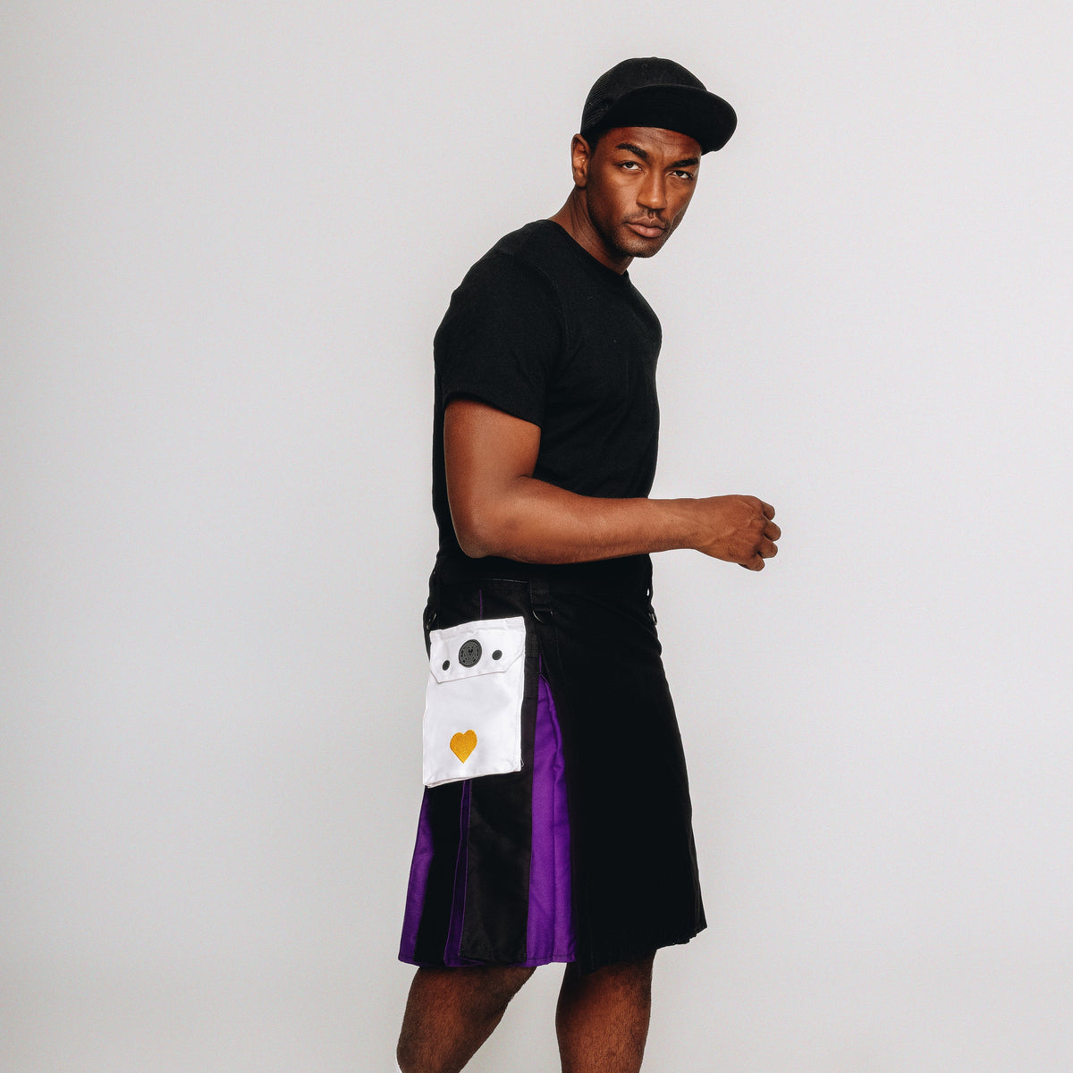 Person wearing a black t-shirt, black kilt with a purple accent, and a cap on a white background