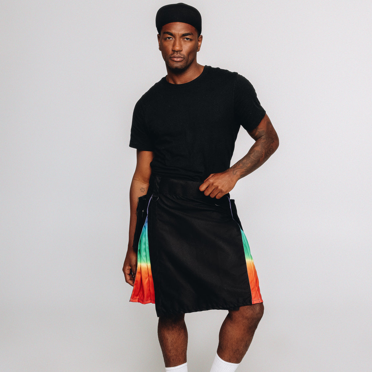 A man modeling a black t-shirt, black kilt with vibrant rainbow-colored side panels, white socks, and black shoes against a plain light gray studio background.
