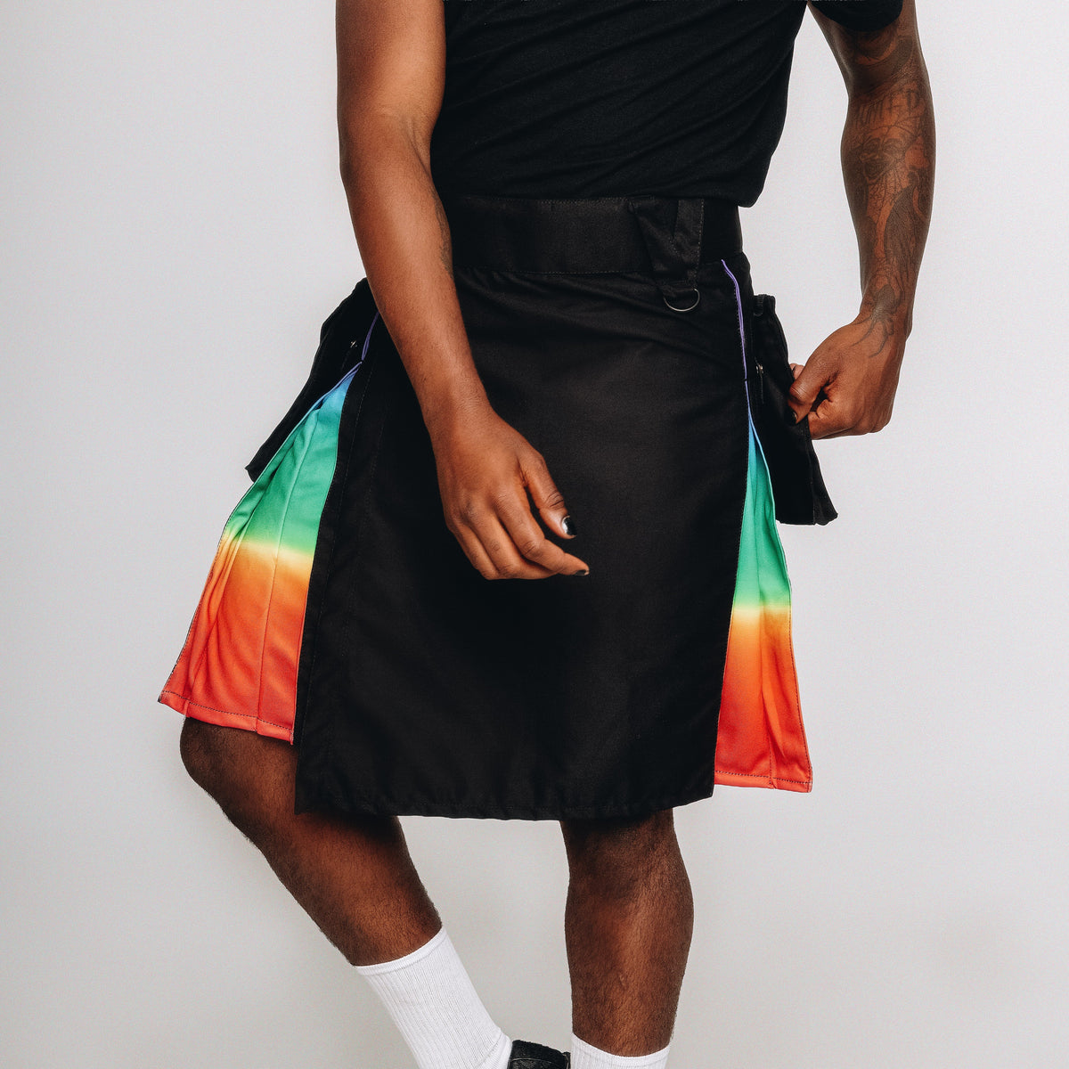 A person modeling a black kilt with vibrant rainbow gradient side panels, styled with white crew socks and black shoes against a plain light gray studio background.