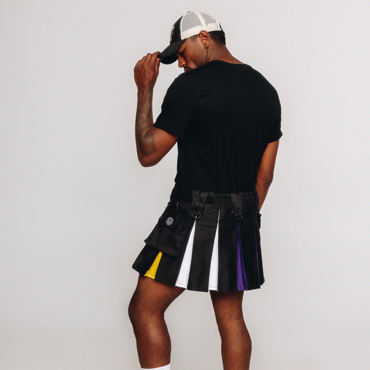 A person wearing a black t-shirt, a black pleated kilt with white, yellow, and purple side panels, and a black and white trucker hat, photographed from the back against a plain light gray studio background.