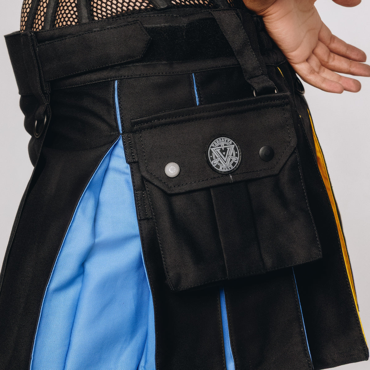 A close-up of a person wearing a black kilt with bright blue side panels and a front utility pocket with a round decorative snap button.