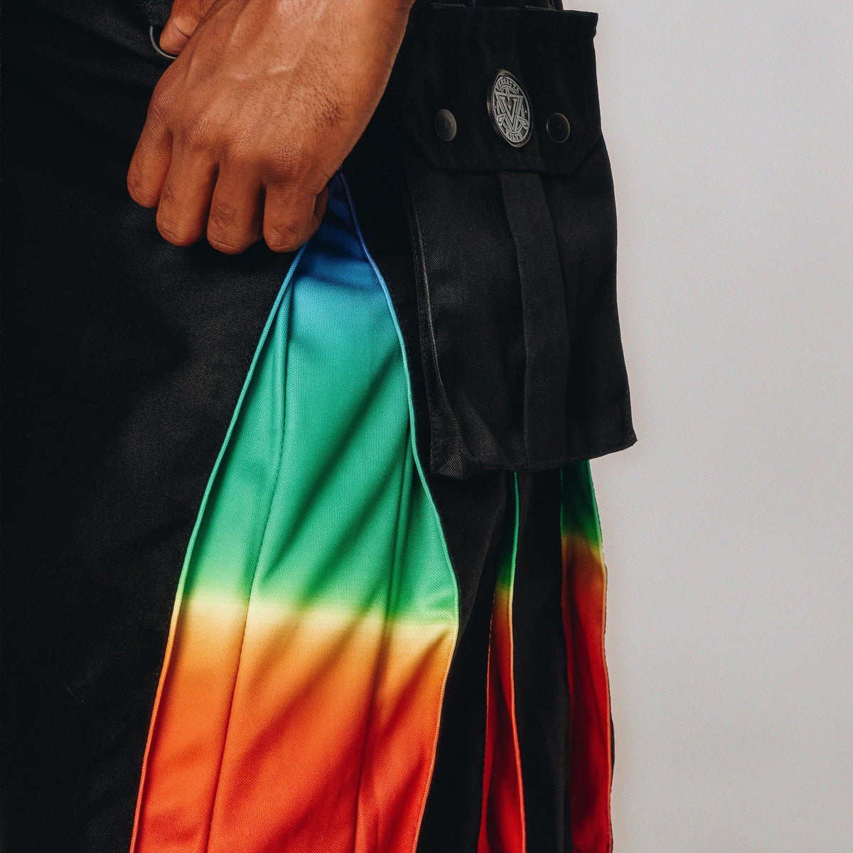 Close-up of a person wearing a black kilt with a prominent rainbow gradient panel in green, blue, yellow, orange, and red, plus a black pouch with metal snaps.