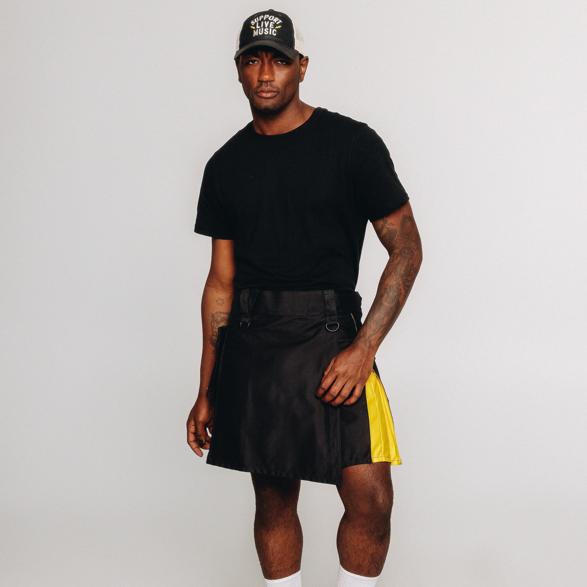 Person wearing a black t-shirt, black kilt with yellow lining, and a black cap on a white background