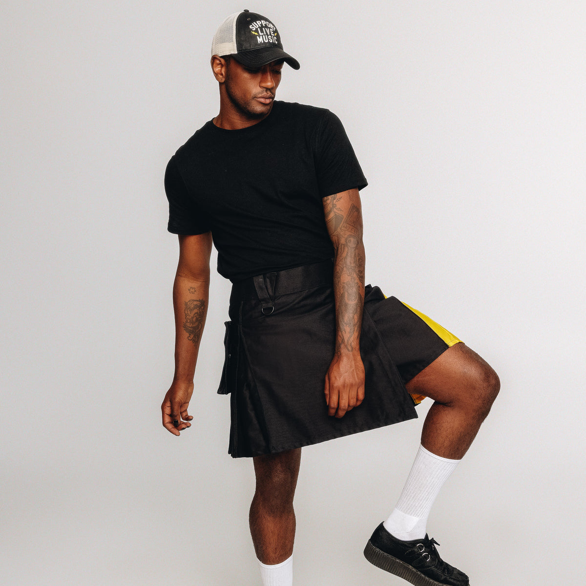 Person wearing a black t-shirt, black kilt, white socks, and black shoes on a plain background