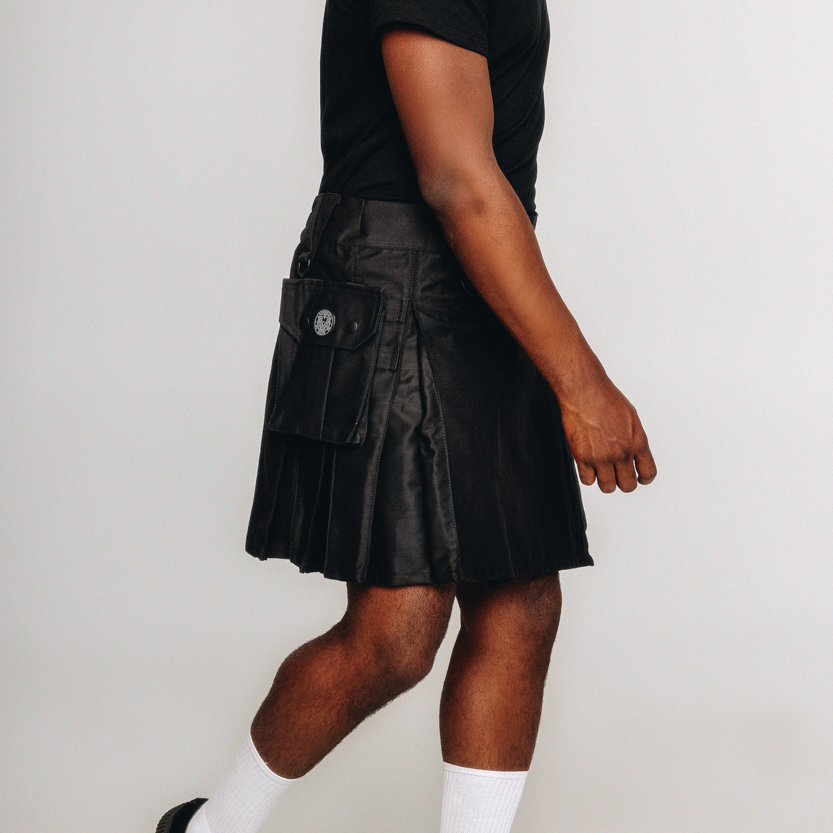 Person wearing a black kilt with white socks and black shoes on a plain background