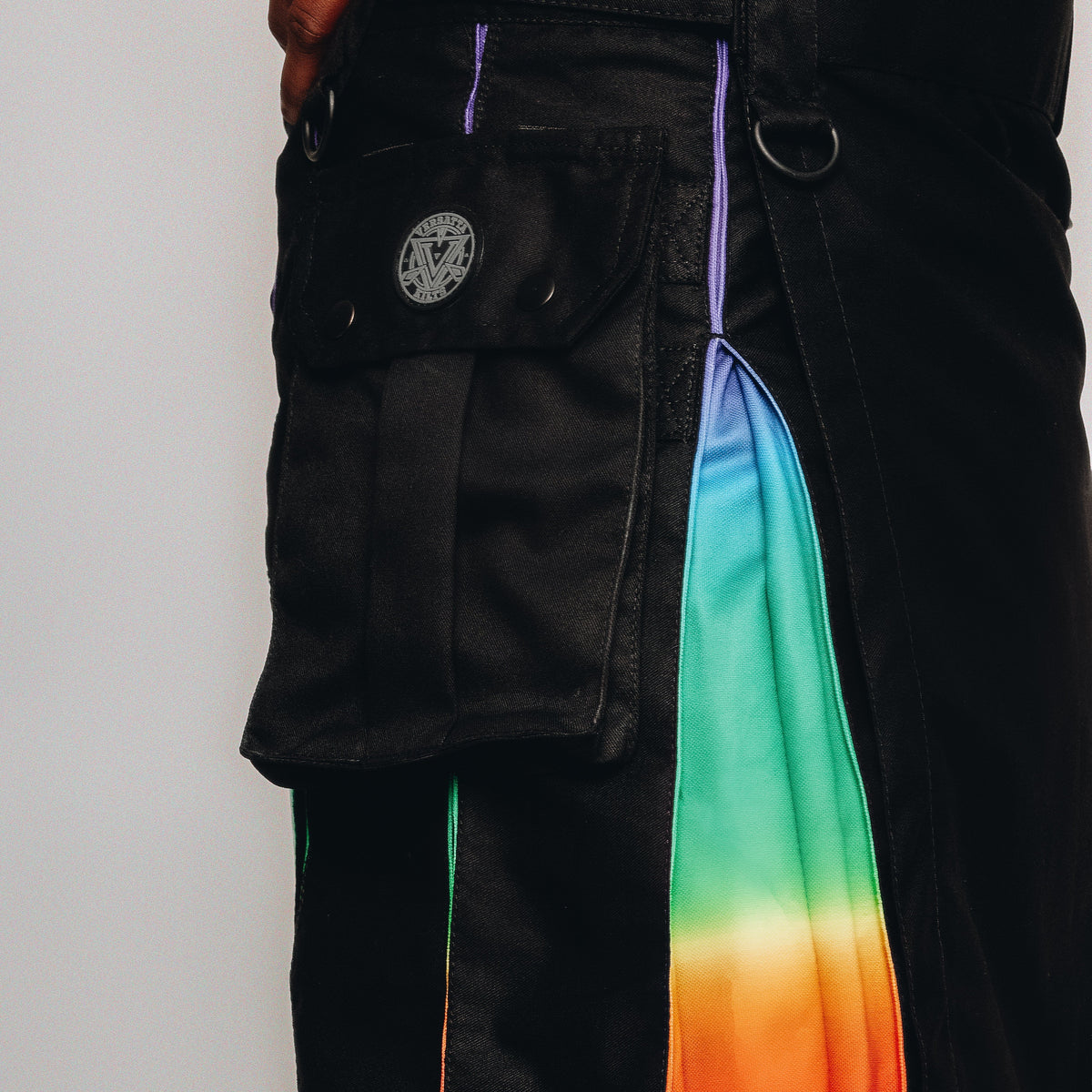 Close-up of a person wearing black cargo-style kilt with a vertical rainbow gradient stripe running down the side and a black belt with a silver-toned buckle.