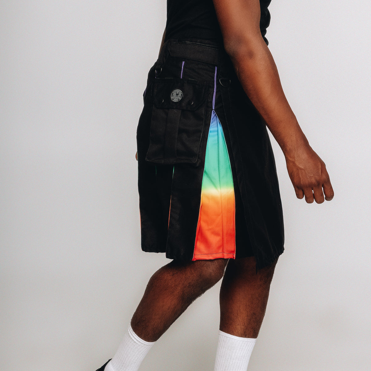 A person wearing black shorts with a vertical rainbow gradient panel on the side, styled with white socks and black shoes against a plain light gray studio background.
