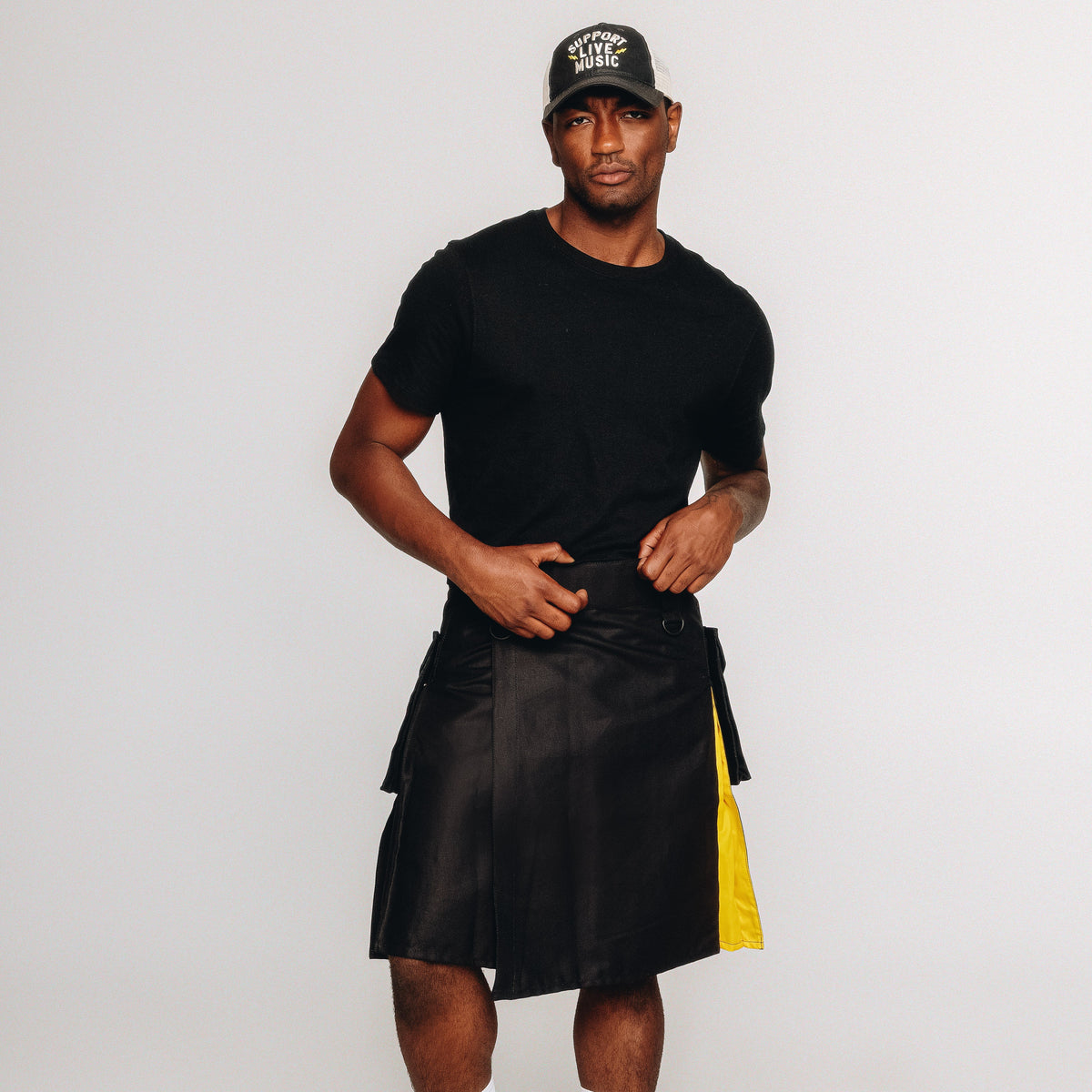 Person wearing a black kilt with nonbinary pride colored pleats and black cap on a white background
