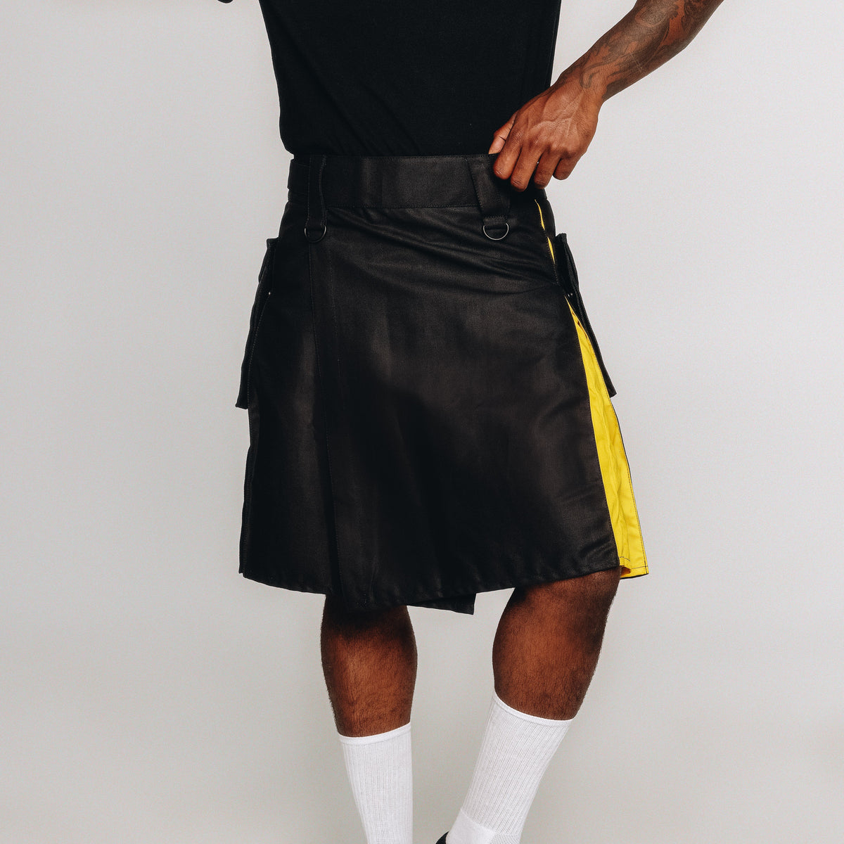 Person wearing a black kilt with nonbinary pride colored pleats, paired with a black top, white socks, and black shoes against a plain light gray studio background.