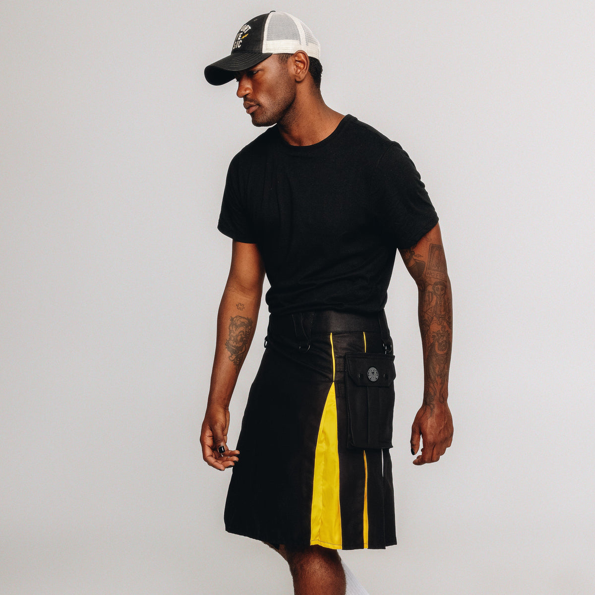 Person wearing a black kilt with nonbinary pride colored pleats and black cap on a white background