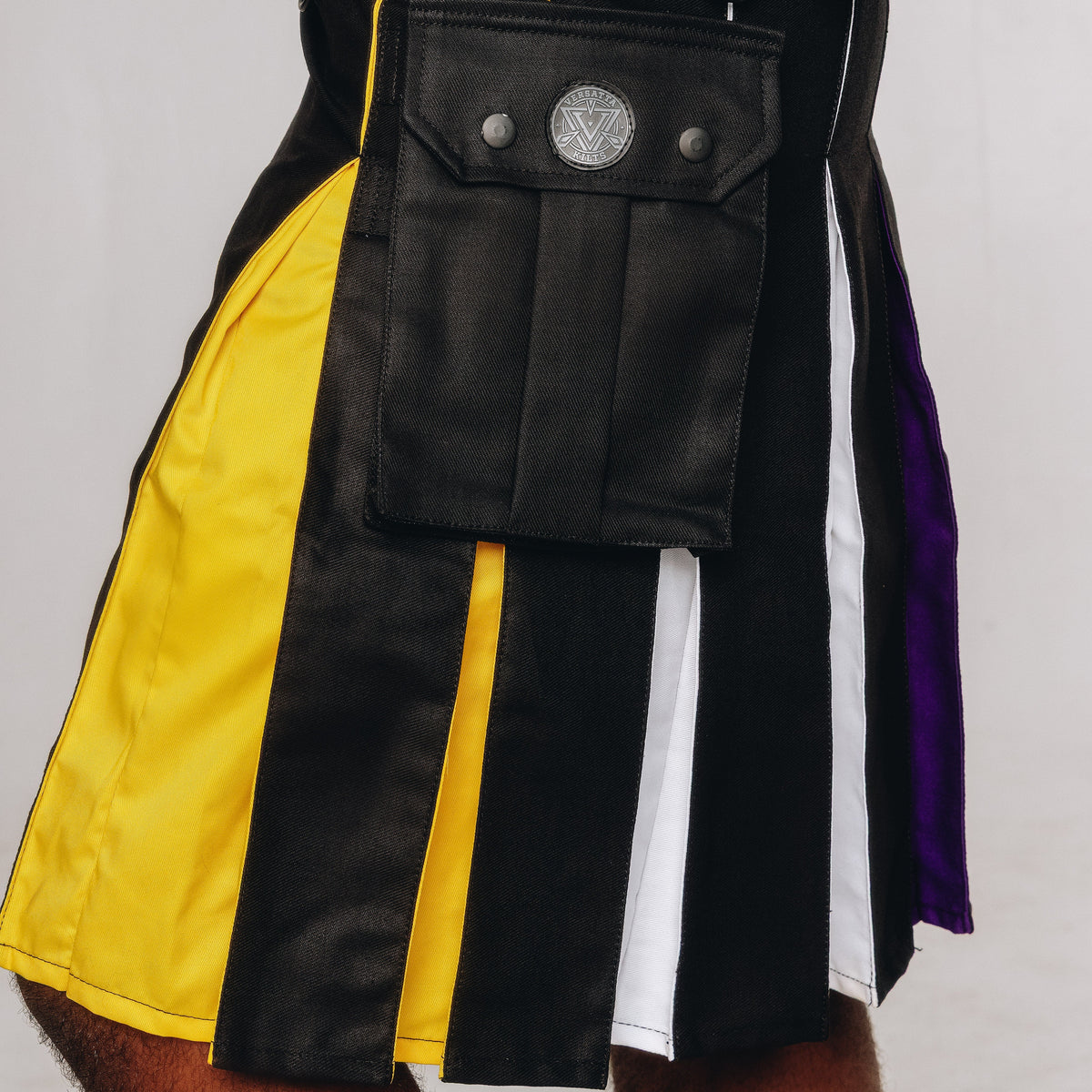 A close-up of a person wearing a black kilt with vertical panels in yellow, white, and purple, plus a large black front pouch pocket with metal snaps.