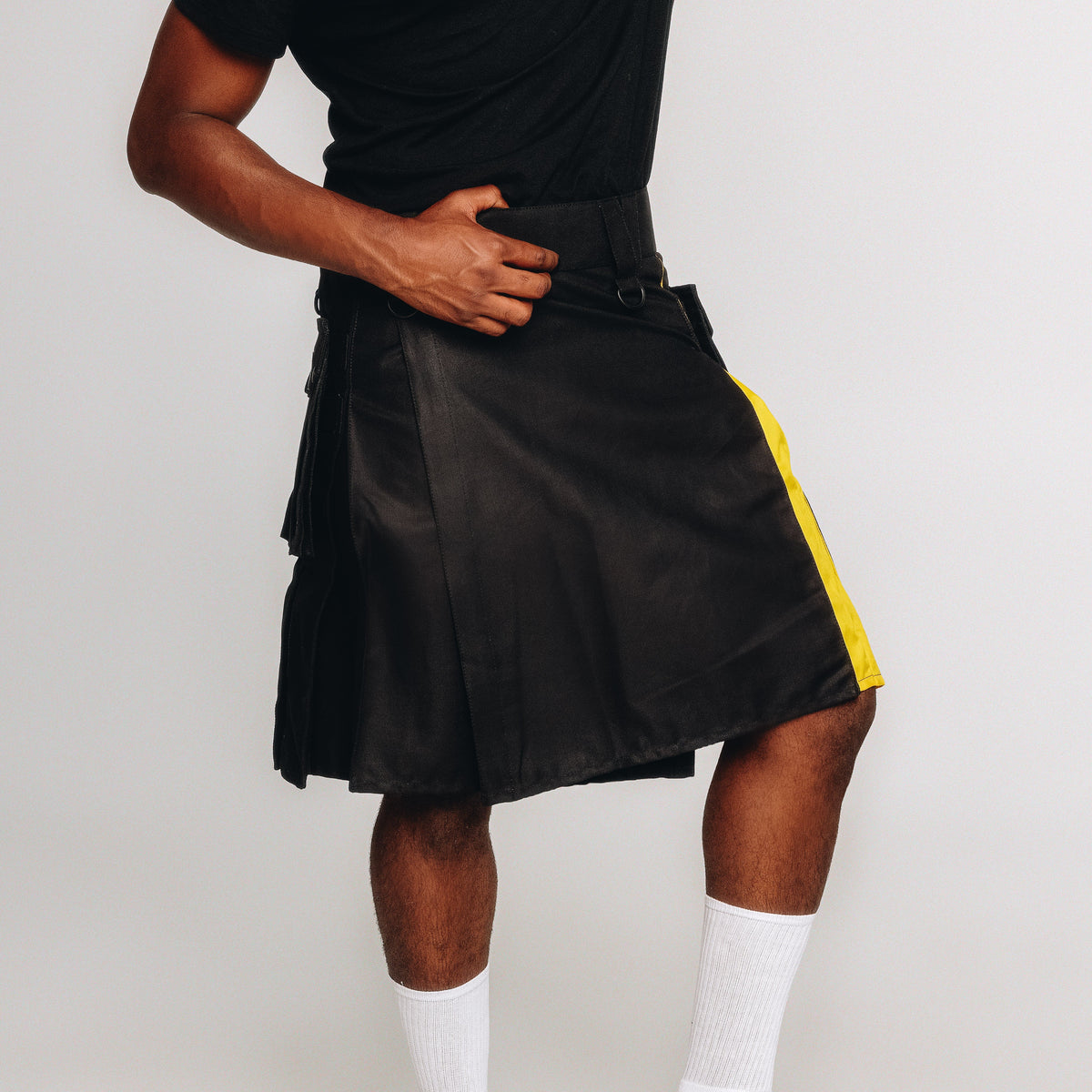 A person wearing a black pleated kilt with a bright yellow side stripe, paired with a black shirt, white socks, and a black belt, photographed against a light gray studio background.