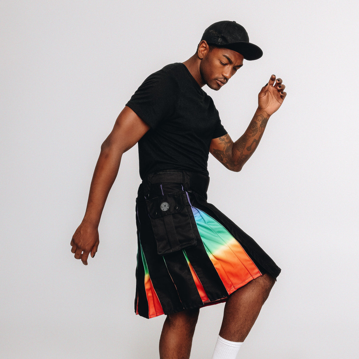 A man modeling a black kilt with a vibrant rainbow gradient panel on the front, paired with a black t-shirt, black cap, and white socks against a light gray studio background.