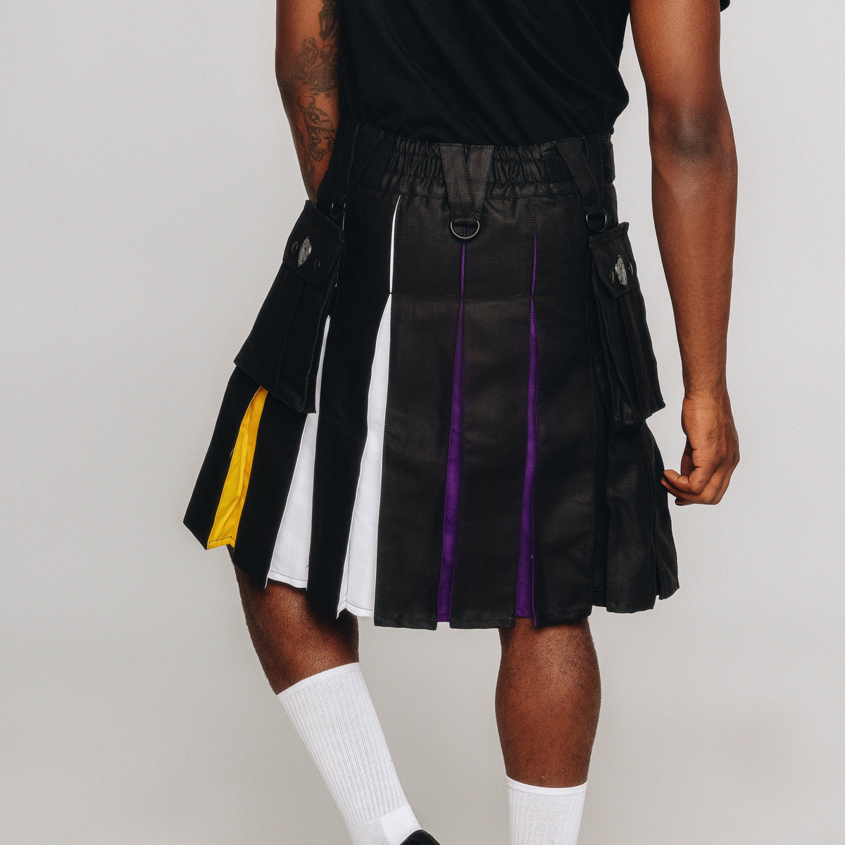 A person wearing a black pleated kilt with vertical panels in white, purple, and yellow, styled with a black shirt, white socks, and black shoes against a light gray studio background.