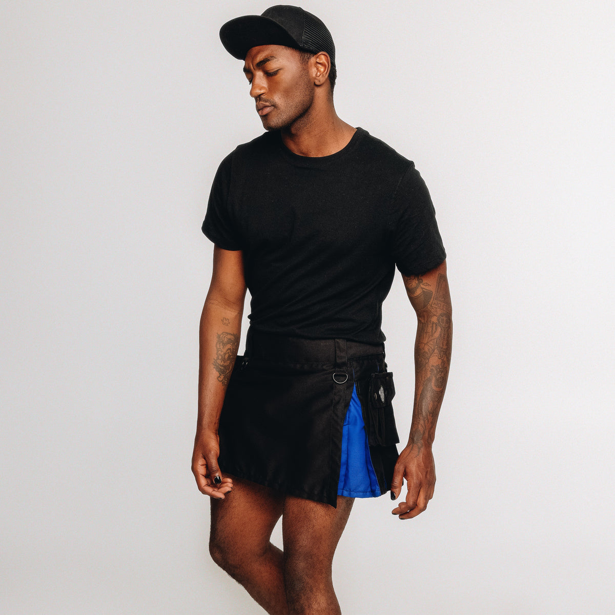 A man wearing a black crewneck t-shirt, a black cap, and a black kilt with a bright blue side panel, standing against a plain light gray studio background.