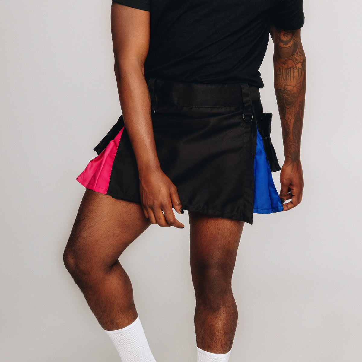 A person wearing a black kilt with bright pink and blue color-block panels on the sides, paired with a black top and white socks, against a plain light gray studio background.