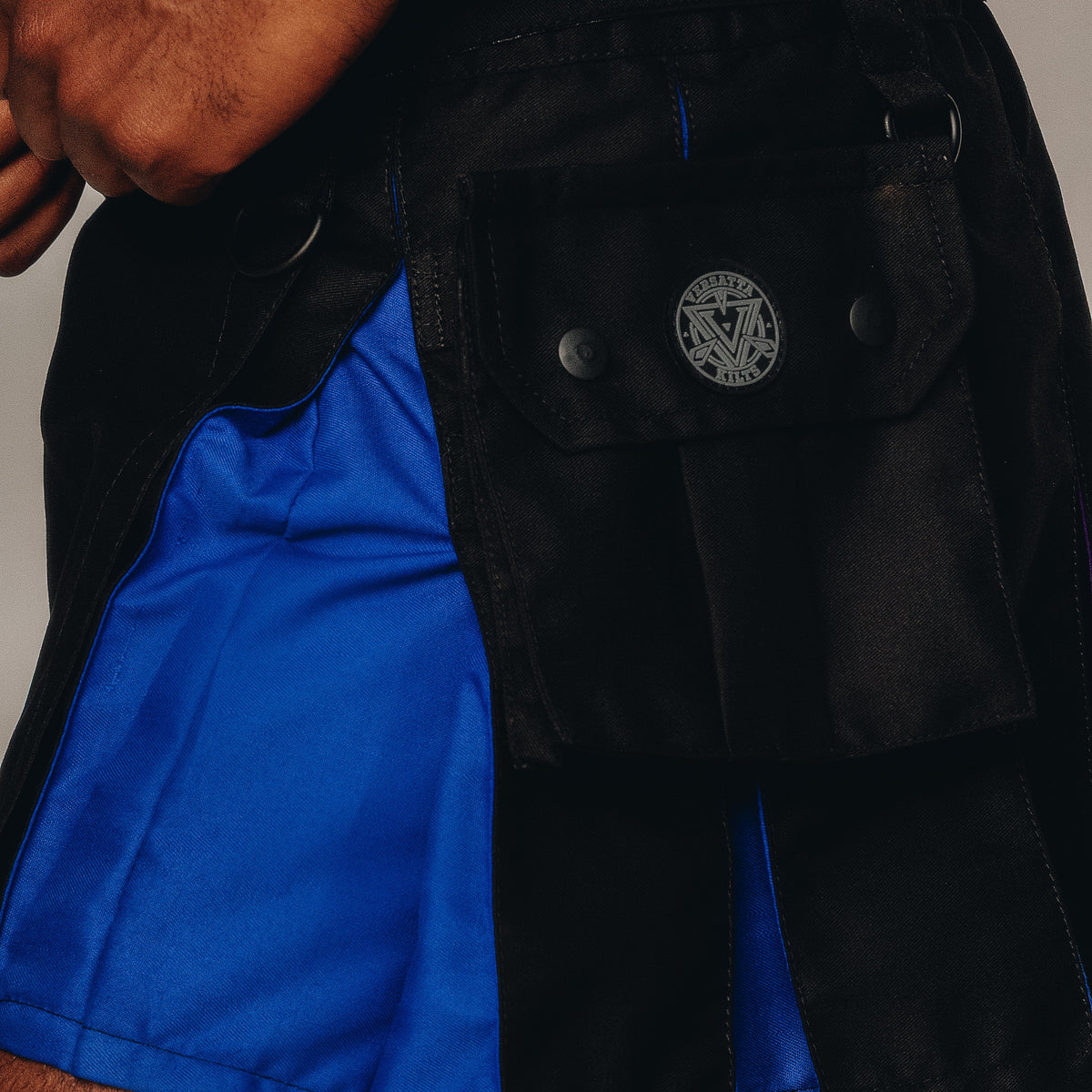 Close-up of black kilt with a bright blue side panel, featuring a flap pocket with a circular metal logo and a visible person's hand.