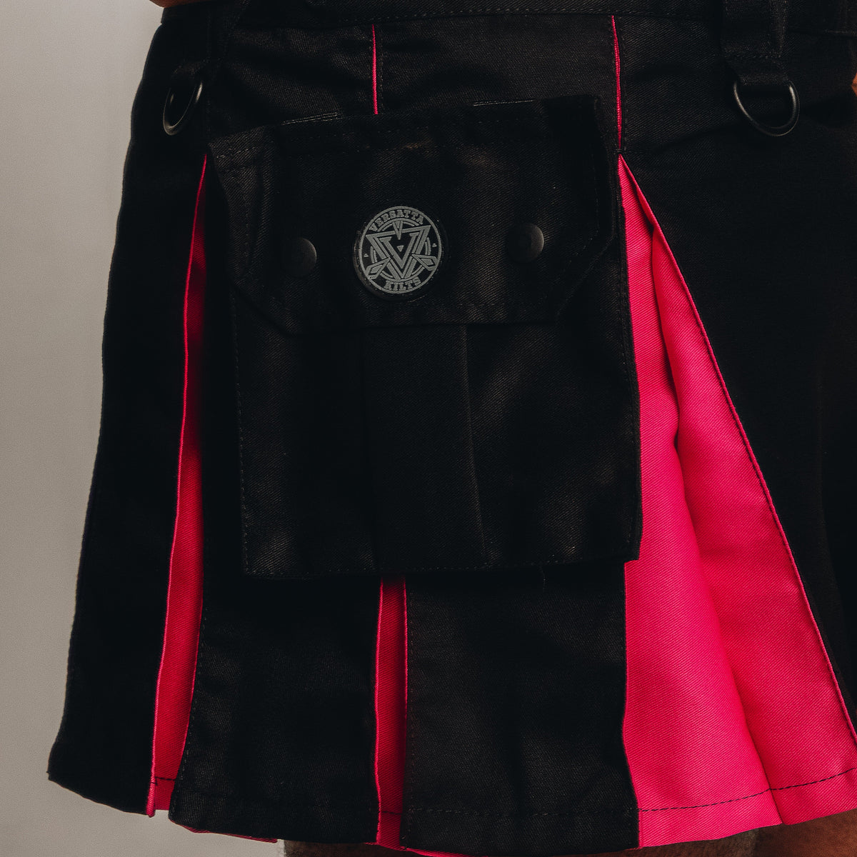 Close-up of a black pleated kilt with bright pink inner panels and a front flap pocket featuring a circular silver-toned emblem.
