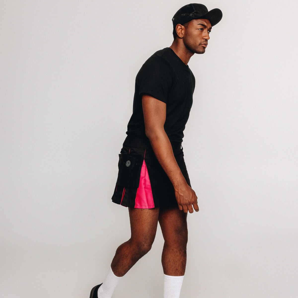 A man in a black t-shirt, black kilt with a bright pink side panel, white socks, and a black cap, shown in profile against a light gray studio background.