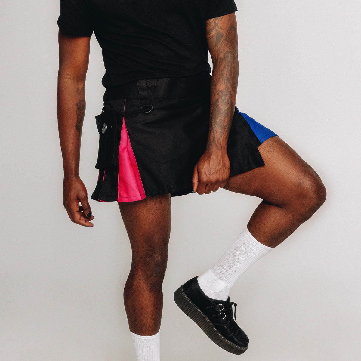 A person modeling a black kilt with bright pink and blue side panels, paired with a black t-shirt, white socks, and black shoes against a plain light gray background.
